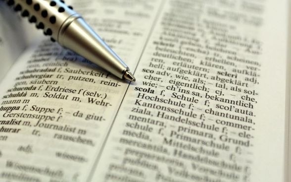Página aberta de um dicionário de romanche com uma caneta apontando para 'scola', palavra em romanche que significa ‘escola’. 