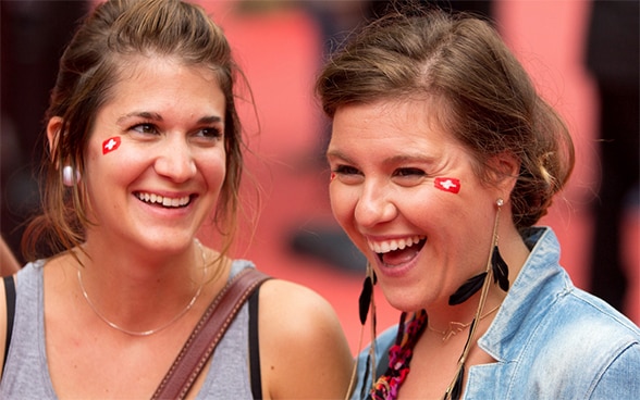 Zwei lachende junge Frauen mit augemalten Schweizer Fahnen auf den Wangen.