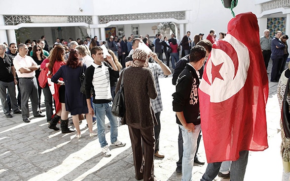 Una lunga fila di persone in attesa davanti a un seggio elettorale in Tunisia. Una persona in primo piano è avvolta in una bandiera nazionale tunisina.