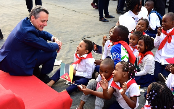 Bundesrat Cassis sitzt auf einem roten Sofa und spricht mit afrikanischen Schulkindern.