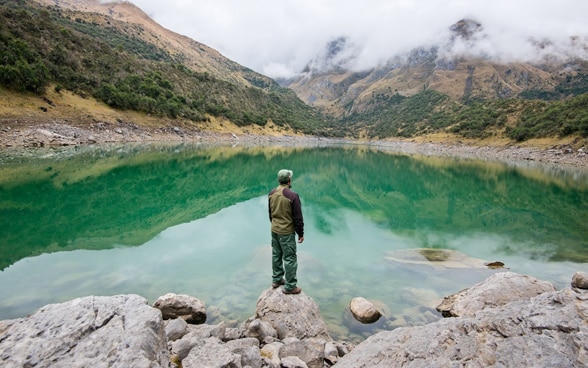  A man standing at the edge of a small lake in a mountainous area.