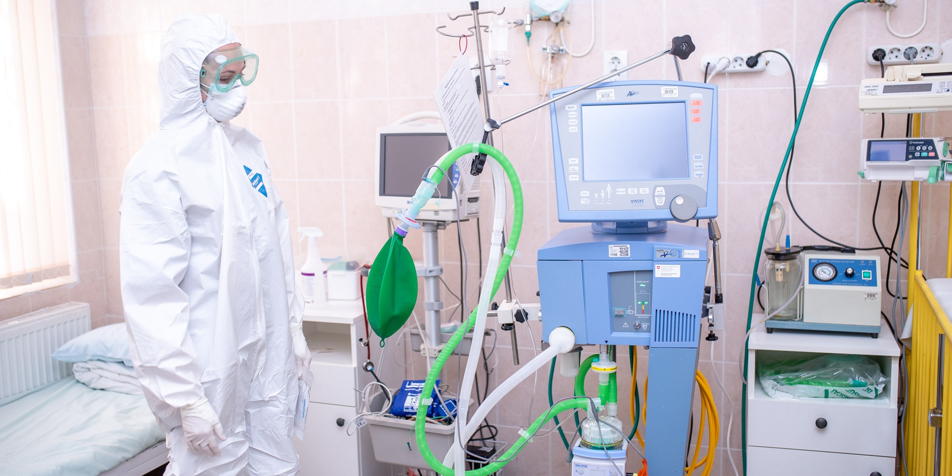 Dans une chambre d’hôpital, une femme portant un masque, des lunettes et une combinaison de protection se tient debout à côté d’appareils médicaux.