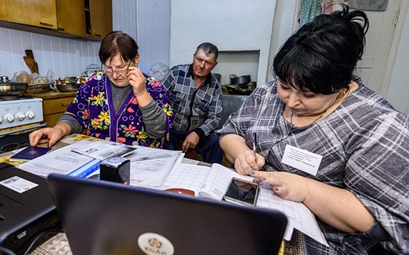  Two women and a man are sitting in a kitchen, working at a computer and reading documents. 