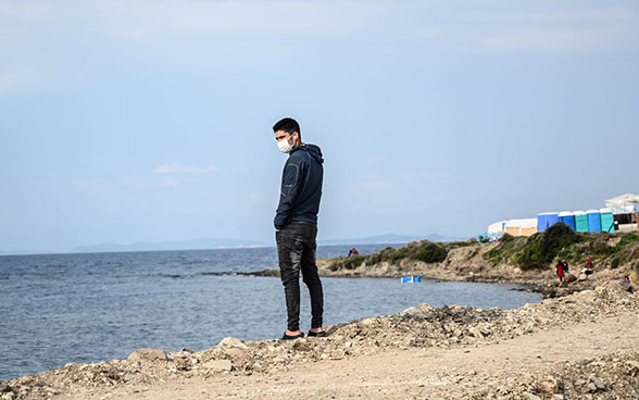 Un migrant debout face à la mer dans le camp de réfugiés de Karatepe, sur l’île de Lesbos, en Grèce, le 29 mars 2021. En haut, à droite, des tentes se dressent sur la côte. En bas, au bord de la mer, se tiennent des femmes vêtues de rouge.