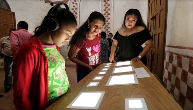 Une petite fille avec des écouteurs regarde des écrans sur une table. A côté d'elle, se trouvent deux femmes.