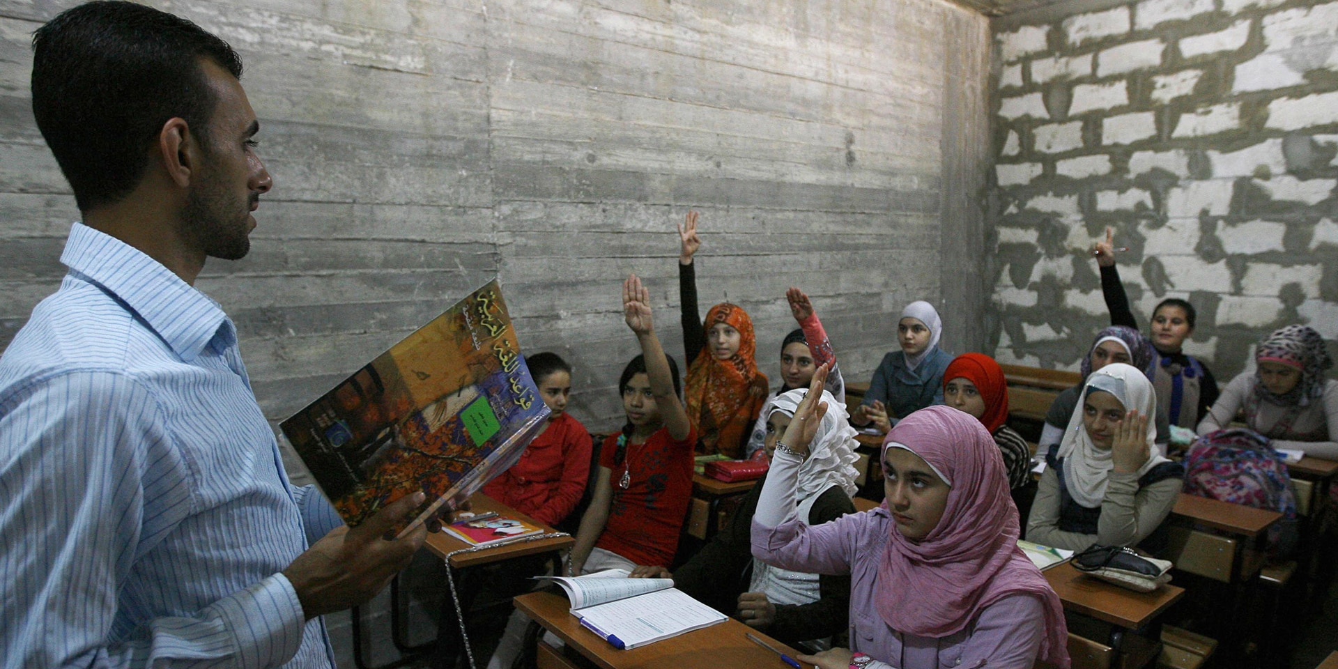 Syrische Flüchtlingsmädchen sitzen in einem Klassenzimmer im Libanon und strecken die Hand.