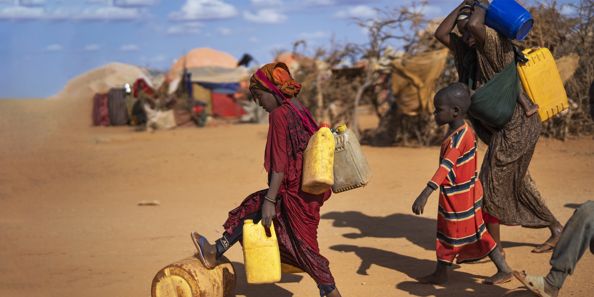 Women and children carry water canisters through a village of simple huts in Somalia.