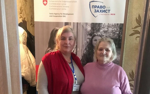 Two older women take each other by the arm and look into the camera.