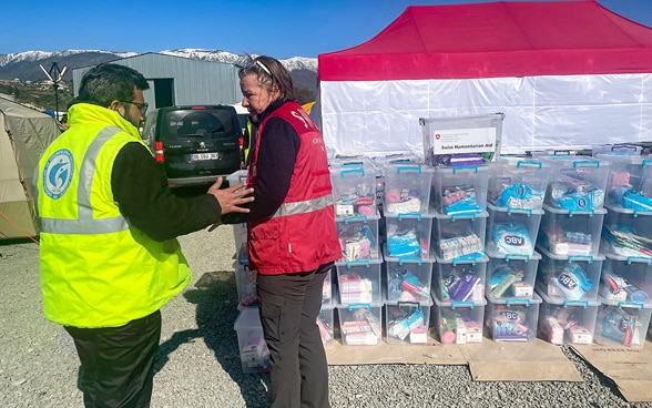 A female member of the Swiss Humanitarian Aid Unit wearing a red waistcoat and a male member wearing a yellow waistcoat stand in front of boxes containing hygiene kits.