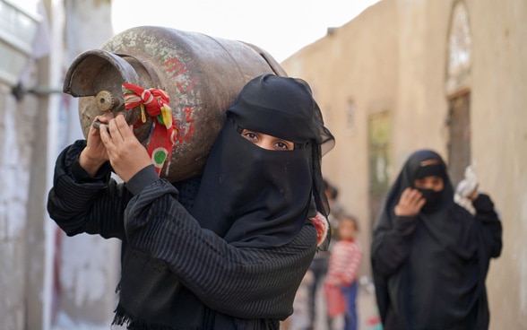 Zwei Frauen tragen eine Gasflasche auf ihrer Schulter und machen sich auf die Suche nach Benzin auf dem Schwarzmarkt.