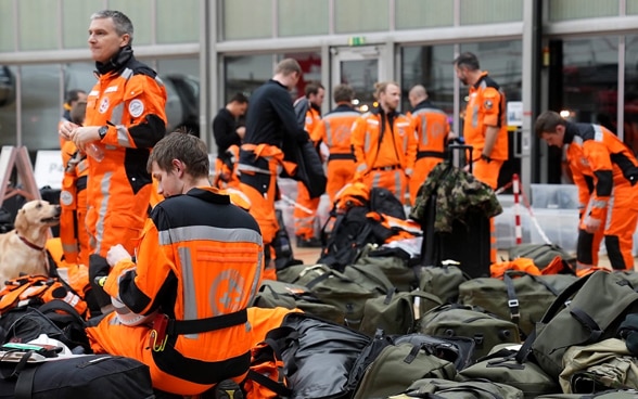 Backpacks and bags are piled up on the floor, with members of Swiss Rescue and a dog standing around them. 