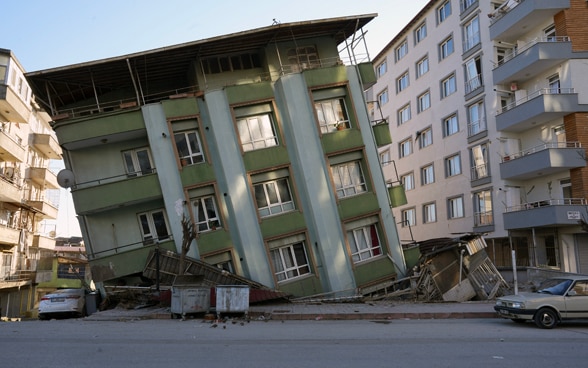 A house stands crooked and tilted on its side between two undamaged houses.