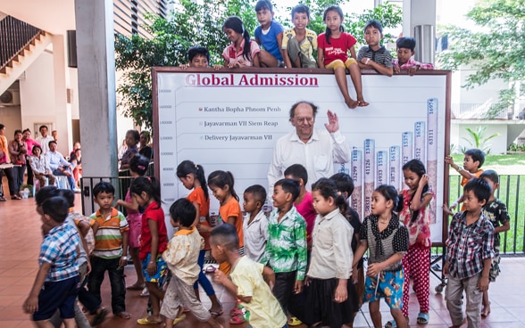Dr Richner surrounded by children in Cambodia in 2013. Behind him is a board with a graph illustrating how many patients come to the hospitals each year. 