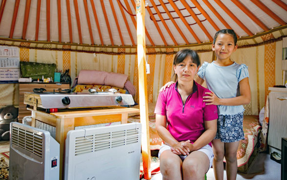 Una madre y su hija están de pie al lado de un nuevo sistema de calefacción moderno en su yurta tradicional, montado con un paquete CHIP.