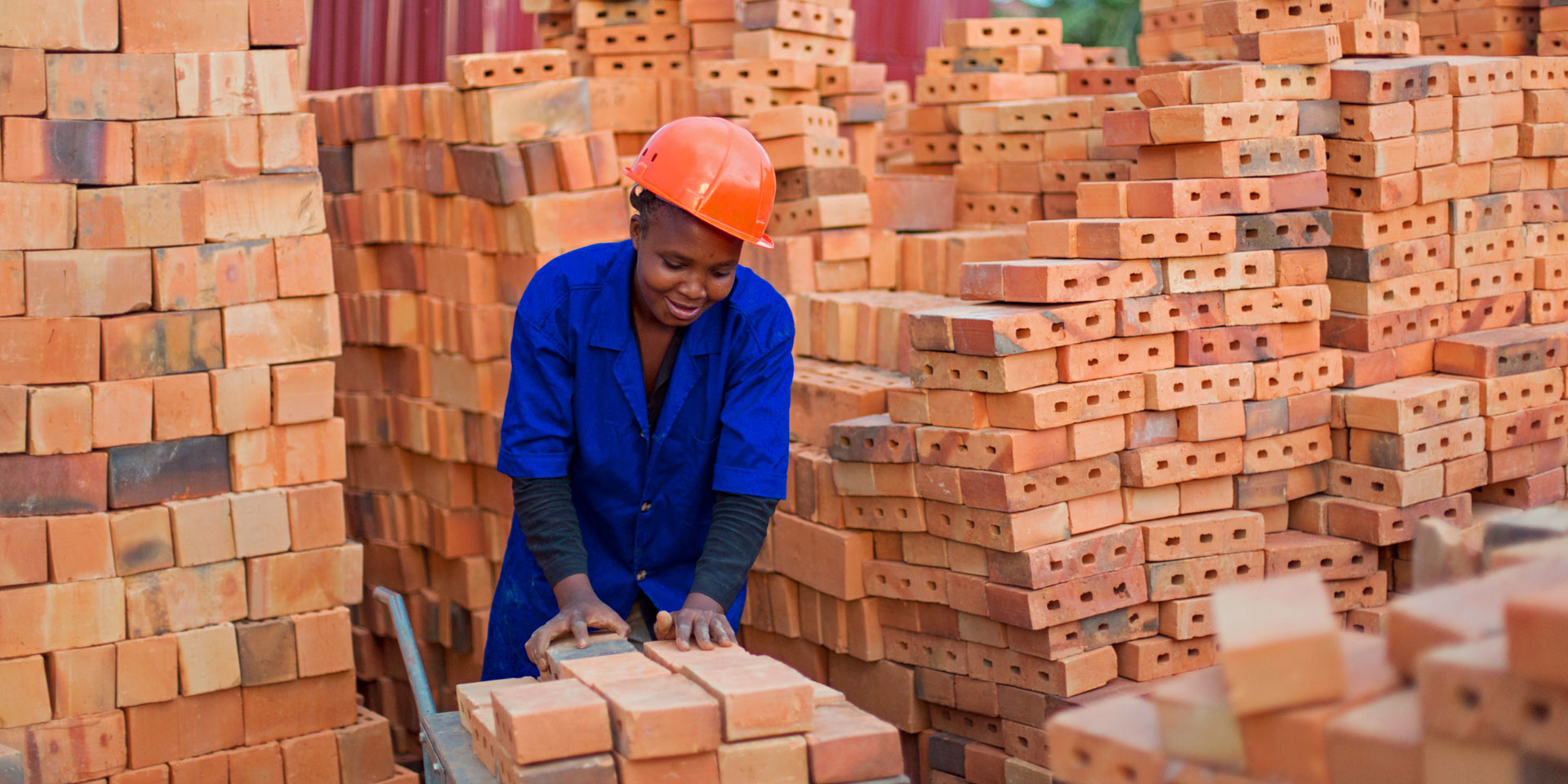 A woman wearing blue overalls and an orange hard hat moves a pile of bricks onto a trolley.