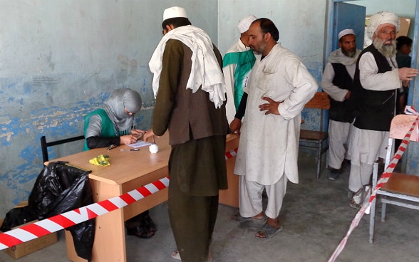 Una donna velata seduta a una scrivania controlla un documento. Diversi uomini attendono il loro turno per votare.  