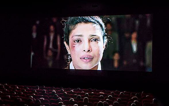 Auf einer Leinwand im Kinosaal ist eine Frau zu sehen. Sie hat eine Verletzung neben dem rechten Auge und weint.