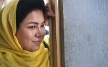 Une femme afghane coiffée du foulard jaune clair traditionnel se tient souriante dans l’embrasure d’une porte.