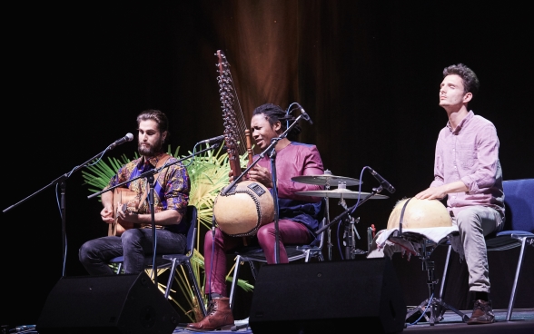 Three musicians with their instruments on stage, sitting in front of microphones. The musician in the middle is singing.