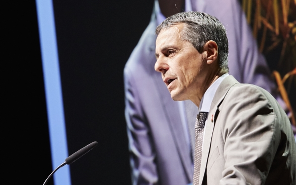 Le conseiller fédéral Ignazio Cassis est debout sur la scène devant le pupitre. Un grand écran se trouve en arrière-plan, sur lequel est projeté le discours.