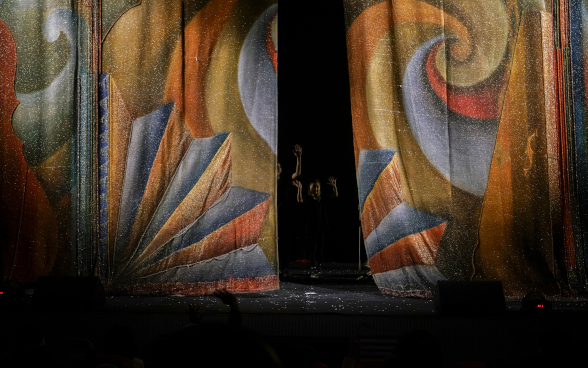 A half-open stage curtain. people can be made out standing behind it in the darkness.