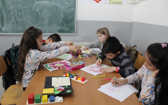 Ragazzi e ragazze fanno disegni colorati seduti in classe intorno a un tavolo di fronte alla lavagna. 