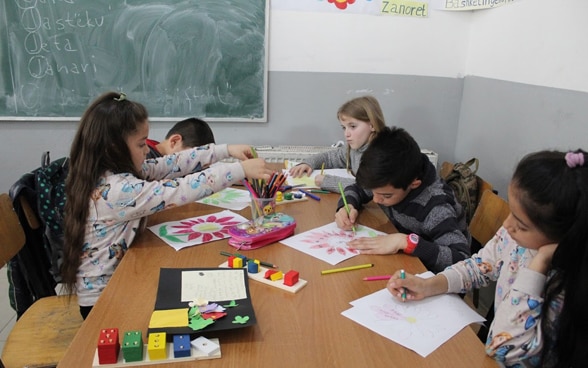 Mädchen und Jungen sitzen im Schulzimmer vor der Wandtafel um einen Tisch und fertigen farbige Zeichnungen an. 