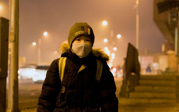 Un bambino con lo zaino in spalla e una mascherina sul volto aspetta lo scuolabus presso una fermata a Ulan Bator.