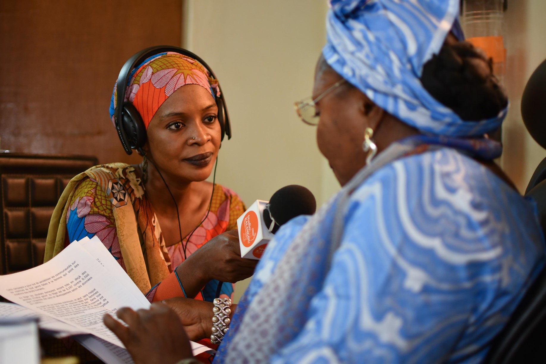  An African woman holds a microphone as she interviews another woman in a radio studio.