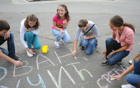 Sechs Jugendliche sitzen im Kreis und schreiben mit Kreide auf den Boden.