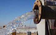 Wasser fliesst aus einem Leitungsrohr. Im Hintergrund sieht man das Flüchtlingslager Azraq