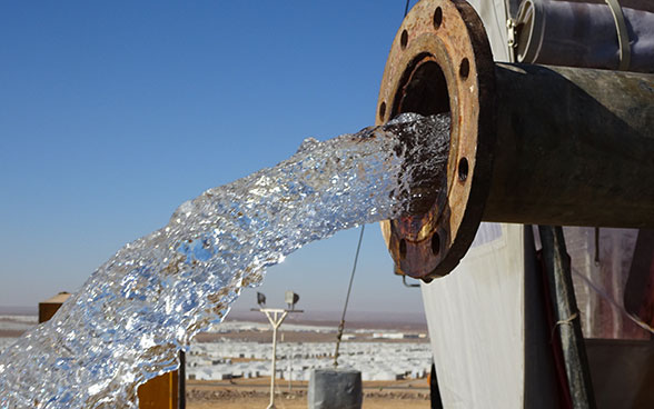 Wasser fliesst aus einem Leitungsrohr. Im Hintergrund sieht man das Flüchtlingslager Azraq
