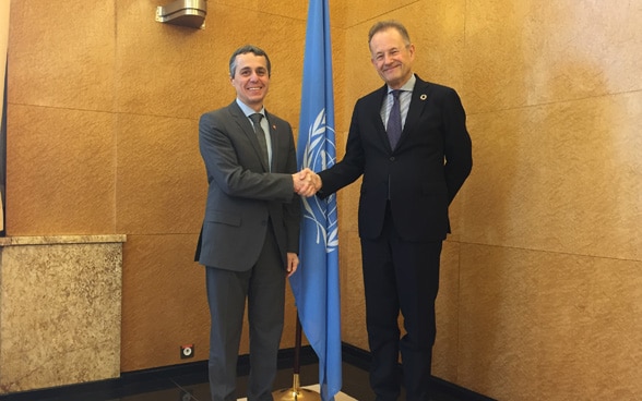 Le conseiller fédéral Ignazio Cassis et Michael Moller, directeur des Nations Unies à Genève, se serrent la main devant un drapeau des Nations Unies.