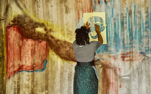 A woman is standing in front of a coloured wall. She is holding a stencil in the shape of a face, which she leans against the wall to paint through the cutouts.