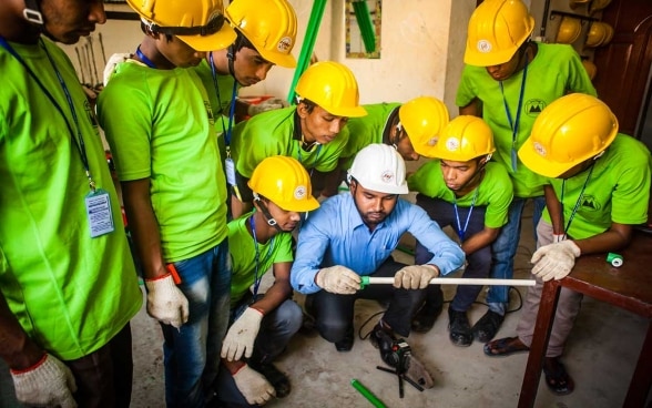 Un groupe d’hommes portant des casques de chantier forment un cercle autour de l’instructeur, qui leur montre comment manipuler des tuyaux en plastique.