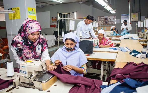 Dans une usine textile, trois couturières travaillent chacune sous l'œil avisé d’une personne chargée de leur formation.