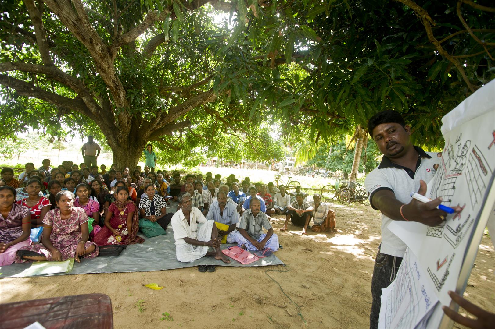 Ein DEZA-Mitarbeiter bespricht mit einer Gruppe Dorfbewohnern ein Bauprojekt. Dutzende Menschen sitzen auf Matten am Boden.