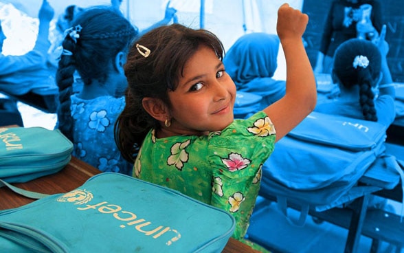 Girls in a classroom put their hands up to answer a teacher's question, while one girl looks back smiling. 