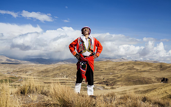Ein nach peruanischer Tradition gekleideter Mann steht auf offenem Gelände im peruanischen Hochland und schaut in den Himmel.