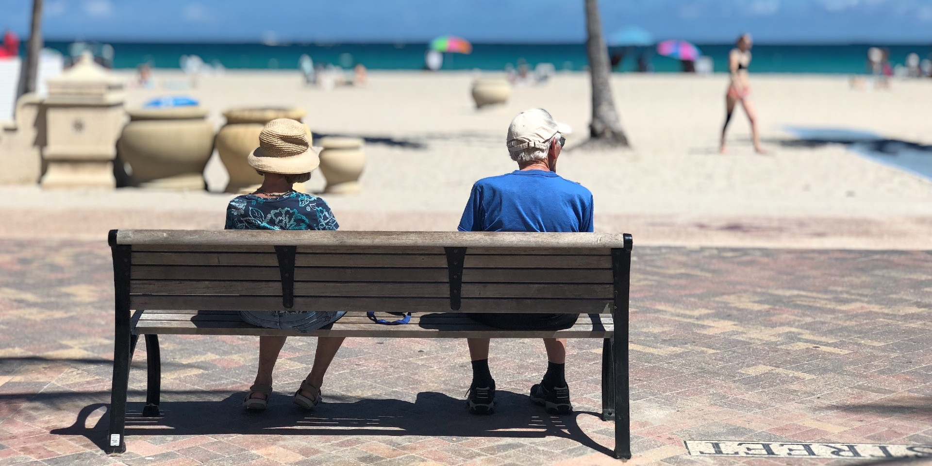 Eine ältere Frau und ein älterer Mann sitzen auf einer Bank und blicken auf den Strand und das Meer.