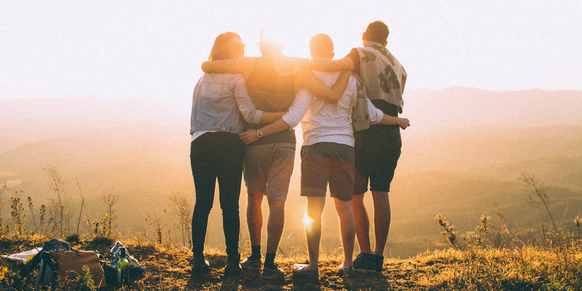 Vier junge Personen umarmen sich und sehen sich auf einem Aussichtspunkt den Sonnenuntergang an.