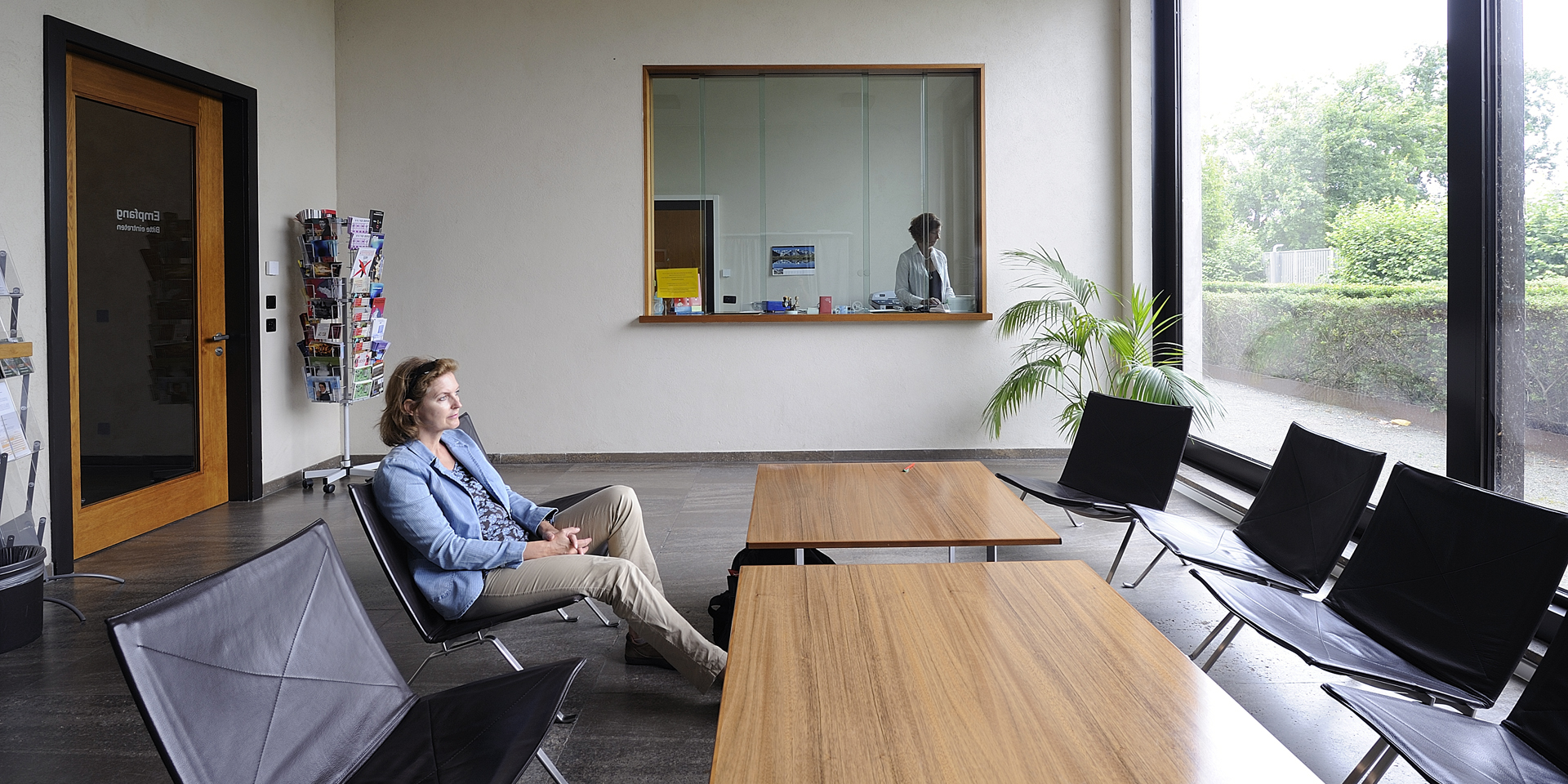 Warteraum des Schweizer Konsulats in Berlin. Eine sitzende Frau wartet, bis sie an der Reihe ist, während ein Mitarbeiter bzw. eine Mitarbeiterin des Konsulats hinter dem Schalter arbeitet.