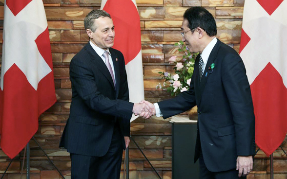 Fumio Kishida et Ignazio Cassis se tendent la main. Les drapeaux du Japon et de la Suisse sont visibles en arrière-plan. 