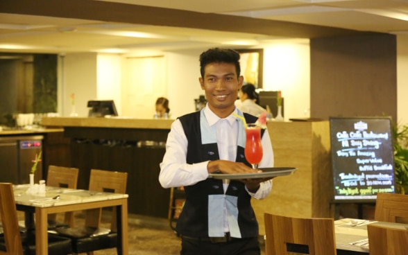 Ein junger Serviceangestellter läuft lächelnd durchs Restaurant mit einem Tablet in der Hand, auf dem ein Cocktail steht.
