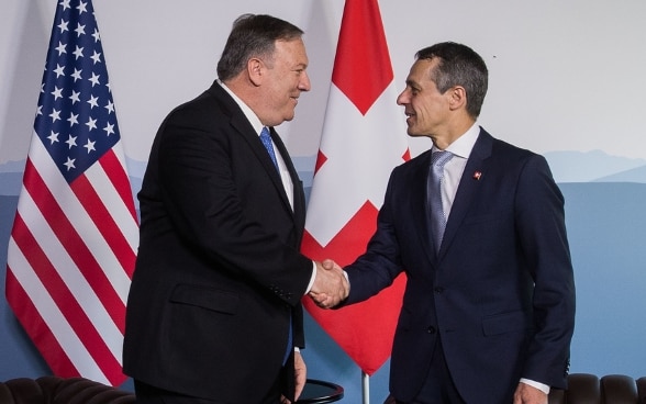 Le conseiller fédéral Cassis et le secrétaire d'Etat américain Pompeo se serrent la main. Les drapeaux de la Suisse et des Etats-Unis sont visibles à l'arrière-plan.