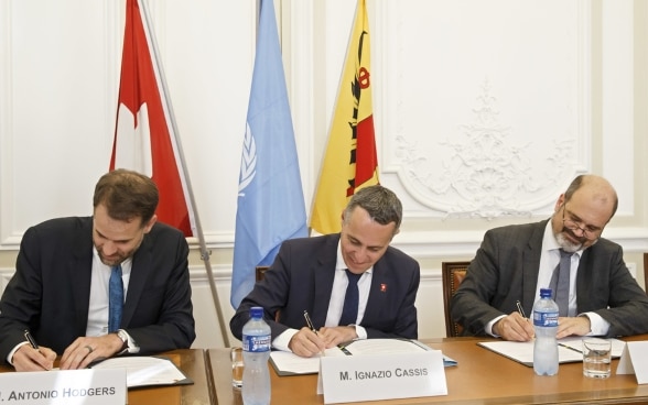 Il consigliere federale Cassis, Antonio Hodgers e Sami Kanaan si siedono a un tavolo di legno e firmano la dichiarazione congiunta.