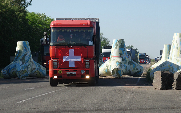 Lorries with Swiss flags drive across large obstacles on a highway.