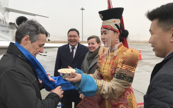 Une femme en costume traditionnel adresse une petite coupelle au conseiller fédéral Cassis, à sa descente d’avion.