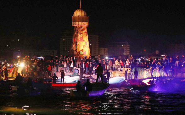 Zahlreiche Fischerboote mit jungen Männern und Frauen versammeln sich vor einem Leuchtturm.