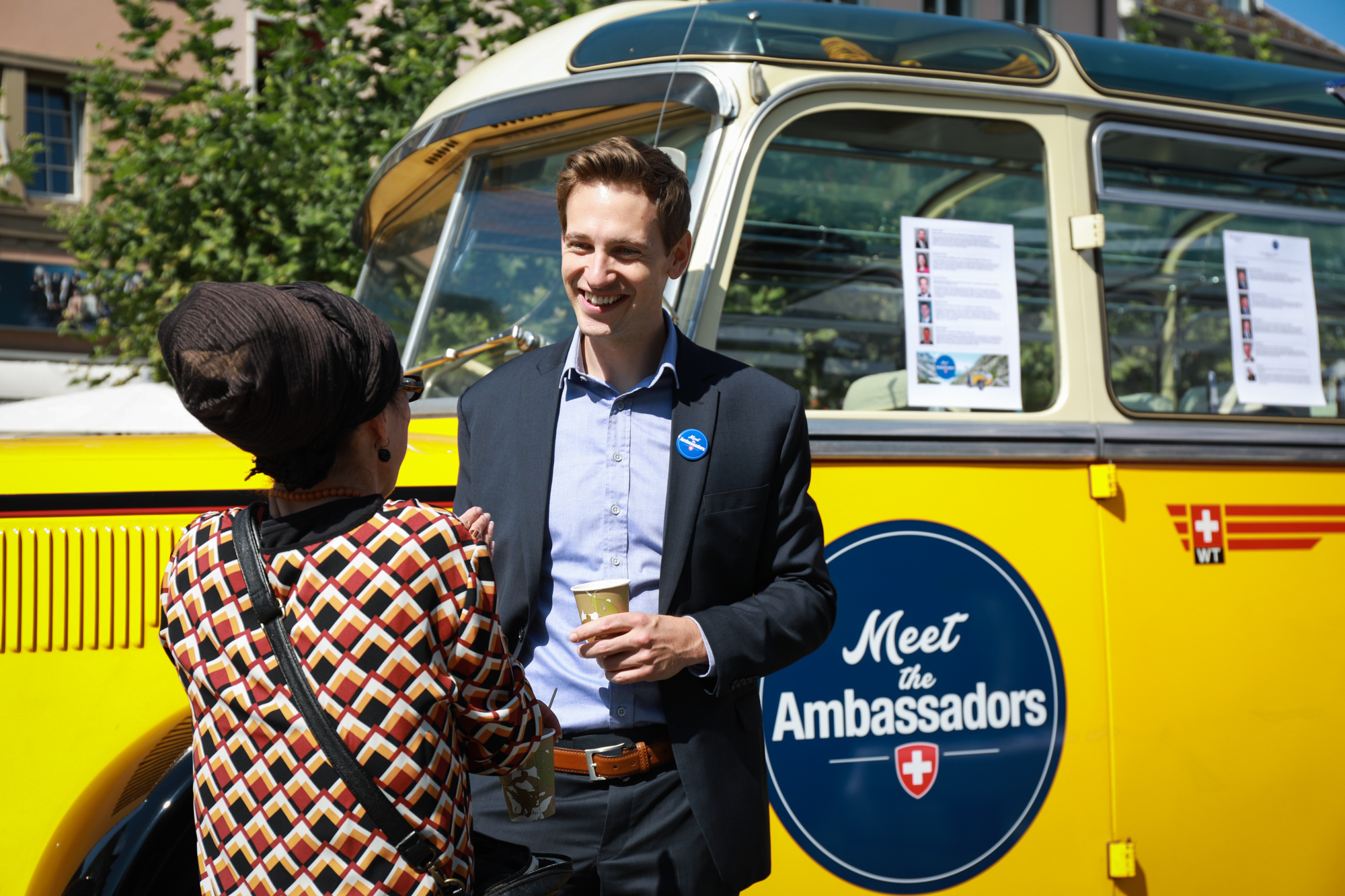 Ein Botschafter unterhält sich mit einem Besucher. Im Hintergrund steht das Postauto Meet the Ambassador